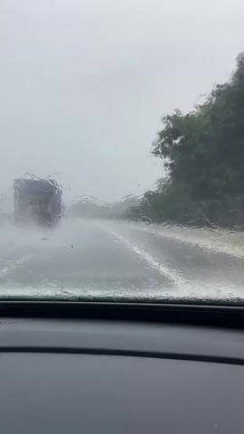 The video shows a view from inside a vehicle traveling on a wet road during a rainstorm. Water is visible on the windshield, distorting the view of the road ahead. A truck is driving in the distance on the same road, also appearing somewhat obscured by the rain. The road has white lane markings. Trees and foliage are visible on the right side of the road, and a fence runs alongside them. The sky is overcast and gray. The dashboard of the vehicle is visible at the bottom of the frame.
