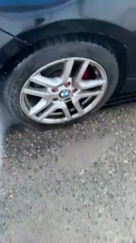 A pressure washer is used to spray the wheel of a car. The car is black, and the wheel has a silver rim and a BMW logo in the center, surrounded by pink accents. Water is being sprayed directly onto the wheel and tire, causing grime and dirt to be washed away. The surrounding pavement is wet and shows signs of previous washing. A drain is visible near the wheel.