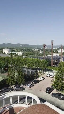 An aerial shot shows a road with parked cars and trees. In the foreground, there is a circular structure with white columns. Beyond the road are tennis courts and more trees. In the background, buildings and hills are visible under a clear sky.