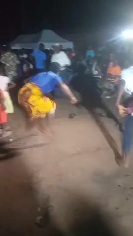The video shows a group of people gathered outdoors at night. Two individuals, one wearing a blue shirt and a yellow skirt and the other a white t-shirt and a maroon skirt, are in the foreground, appearing to be engaged in some form of dance or ritual. They are barefoot and their movements are energetic, with dust being kicked up around them. In the background, other people are visible, some standing and watching, and some sitting. There are tents and bright lights illuminating the scene. The ground appears to be dirt or sand. The overall atmosphere suggests a lively outdoor gathering or performance.