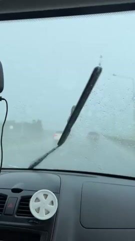 The video shows a view from inside a car during a rainstorm. Raindrops are visible on the windshield, and the windshield wipers are in motion, clearing the view of the road ahead. Other vehicles can be seen on the road in the distance, shrouded in the rain and fog. The dashboard of the car is visible in the foreground, with a white air freshener on the vent. The overall environment is overcast and wet due to the rain.