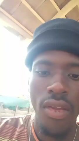 A young Black man wearing a black knit cap, a horizontally striped brown, orange, and white shirt, and a silver cross necklace is recording a selfie video outdoors. He smiles and sticks out his tongue at the camera. The camera angle shifts to capture the sky, trees, and the corner of a building in the background. The sky is partially cloudy, and a palm tree is visible. The man is standing near a wooden structure. He is lip-synching to the song playing in the background.