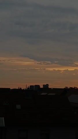 The video shows a sunset over a cityscape. The sky is filled with dramatic clouds, ranging from dark gray at the top to golden orange near the horizon, where the sun is setting. Beams of sunlight are piercing through the clouds, creating crepuscular rays. In the foreground, dark silhouettes of buildings and rooftops are visible. Some rooftops have solar panels. The overall atmosphere is serene and awe-inspiring.