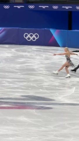 A man and woman figure skate on an ice rink in front of a banner reading "MILANO CORTINA 2026" and displaying the Olympic rings. The woman is wearing a silver dress with a black design. The man is in a black costume. Both have dark hair. They separate, performing their own routines to the music. There are spectators in the stands. A videographer is taking footage.