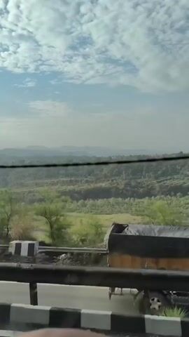 A video taken from inside a vehicle, looking out a window, shows a view of rolling hills and a cloudy sky. A road with a guardrail is visible in the foreground. A white car is parked on the side of the road.
