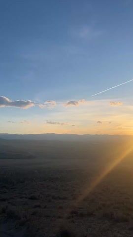 The video shows a wide landscape view at sunset. The sun, obscured but beaming brightly, casts long rays across a dark, low-lying field of shrubs and plants. In the distance, there is a hazy outline of hills or mountains. The sky is a mix of blue and orange hues, with light clouds scattered around, some tinged with a rosy color.  A long white contrail stretches across the sky, likely left by an airplane. The overall impression is one of vastness and a peaceful, natural setting.