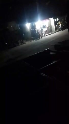 The video, filmed in the dark, appears to show a street scene at night. A building with bright lights is visible in the background, possibly a store or restaurant. Several people are standing near the building. The foreground is mostly dark, with some indistinct shapes that could be furniture or other objects. The camera is stationary.