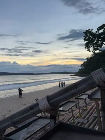 A beach scene at dusk, with a gentle ocean and a cloudy sky. Several people are visible on the sand and in the shallow water. A wooden structure with rope railings is in the foreground, with what appear to be empty tables. The lighting suggests either early morning or late evening.