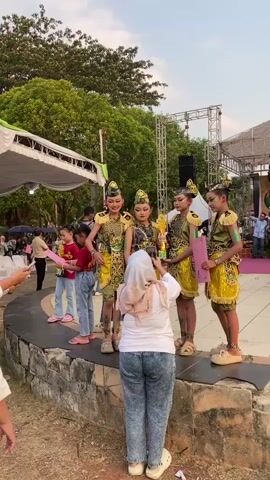 A group of four young females in ornate yellow and gold costumes are posing on a stage for a photograph. One of the girls is holding a trophy and a pink certificate. A woman in a hijab and jeans is photographing the group with a mobile phone. In the background, there are trees, a stage with lighting equipment and a banner, and other people standing and watching. The overall setting appears to be an outdoor event.