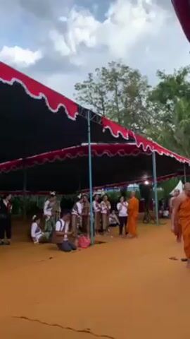 Here is a concise description of the video:

The video shows a group of traditionally dressed young women standing under a large tent with a red and white scalloped edge, along with a few other people. One man is kneeling with a camera, seemingly taking photos. Two Buddhist monks in orange robes are present, and one is interacting with the group of women. The setting appears to be outdoors, with trees visible in the background. The ground is covered with what looks like orange carpet or material. A loudspeaker sits next to the group of women.