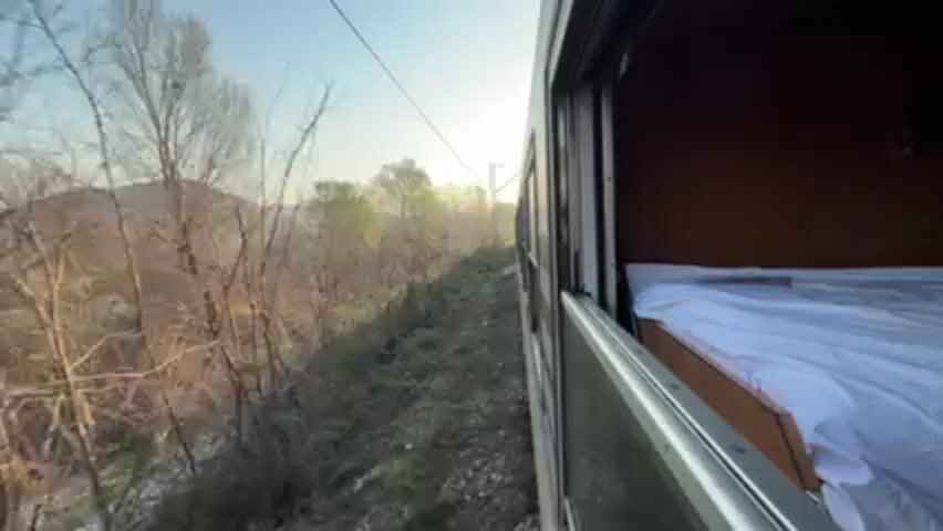 The video shows a view from a train window. Outside the train, a rural landscape is visible with trees and a distant hill under a clear blue sky. A utility wire stretches across the top of the frame. Inside the train car, a bed with white linens is visible. The train is moving, as indicated by the changing scenery.