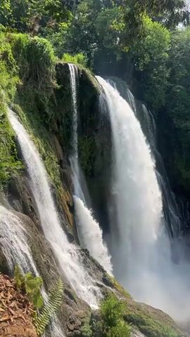 The video shows a set of powerful waterfalls cascading down a lush, green hillside. Waterfalls of varying sizes are visible, with the largest one creating a significant mist at the base. The surrounding environment is densely vegetated with trees, ferns, and other greenery. The landscape consists of moss-covered rocks and soil. The sky is partially visible at the top of the frame, showing some scattered clouds and sunlight filtering through the trees. The overall atmosphere is one of natural beauty and powerful natural forces.