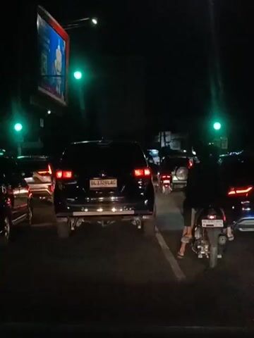 The video shows a traffic jam at night. There are several cars and motorcycles on the road. The traffic lights are green. There is a large billboard on the left side of the screen with a picture on it. A person is riding a motorcycle in the right lane. The road appears to be wet.