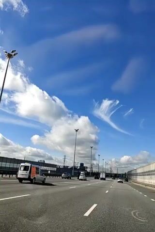 The video shows a view from a vehicle driving on a highway. The sky is blue with scattered white clouds, some of which are wispy and feathery, while others are large and puffy. The highway has multiple lanes with traffic moving in the same direction. Several vehicles are visible, including cars and a white van with an orange stripe on the back. Tall lampposts line the sides of the highway, and there are sound barriers or walls on the right side. On the left side, there are industrial structures and power lines. The video appears to be a continuous shot from the perspective of someone in the moving vehicle.