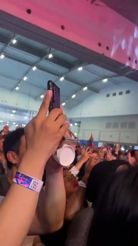 The video shows a crowd of people at a concert, with many holding up their phones to record the performer on stage. The stage is lit with bright red and white lights, and the performer is singing into a microphone. The crowd seems enthusiastic and engaged, and they are all facing the stage. The venue appears to be a large, indoor space with a high ceiling and lighting fixtures.