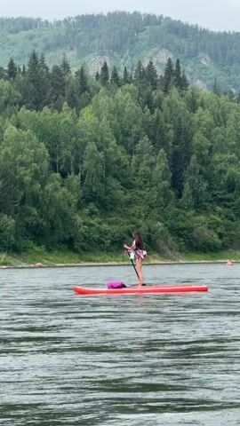 The video shows a young woman paddleboarding on a wide, calm river. She is standing on a red paddleboard with a pink bag behind her. She is holding a paddle and appears to be moving forward, though the movement is slow. The woman is wearing a patterned top and shorts. She has long, dark hair.

The setting is a natural landscape with a dense forest covering rolling hills and mountains in the background. The trees are lush and green, indicating a summer or late spring season. The sky is overcast, suggesting a cloudy or potentially rainy day. The water appears slightly rippled, but overall calm. There are faint orange buoys visible in the distance on the water, and a blurred figure on another watercraft.

The activity is recreational paddleboarding, a peaceful and solitary activity enjoyed on the water. The overall mood is serene and tranquil, fitting with the natural environment and the slow pace of the paddleboarding. No text is visible in the video. The scene is likely taking place in the afternoon due to the diffused light. The location context suggests this is in Ust-Mana, Russia.