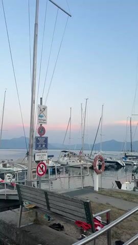 A video shows a marina filled with sailboats. The sky is a soft blue with hints of pink from the setting sun. Mountains are visible in the distance. In the foreground, a wooden bench sits on a paved area, with some belongings scattered around it, including a red shirt and black sandals. A life preserver is visible on a pole near the water. The boats are docked and appear to be at rest. The overall atmosphere is peaceful and serene.