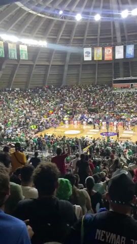 The video shows a basketball game taking place in a large, crowded arena. The perspective is from the stands, looking down onto the court. The court is illuminated, and there are players on the court, some wearing yellow and others green uniforms. The crowd is large and diverse, with many spectators wearing green clothing. There are banners hanging from the ceiling of the arena. A large screen above the court displays the text "¡VAMOS, TRIBU!". The game appears to be in progress, with spectators cheering and reacting. The overall atmosphere is energetic and lively.