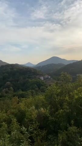 The video shows a drone shot from a high vantage point, overlooking a lush green valley with rolling hills and mountains in the distance. The sky is partly cloudy with the sun shining brightly, casting a warm glow over the landscape. There are a few scattered houses nestled among the trees, suggesting a rural or village setting. As the drone moves, the view shifts, revealing different perspectives of the valley. The overall impression is one of natural beauty and tranquility, with a touch of human presence in the form of the houses. The video appears to be a landscape or travel video, designed to showcase the scenic beauty of the area.