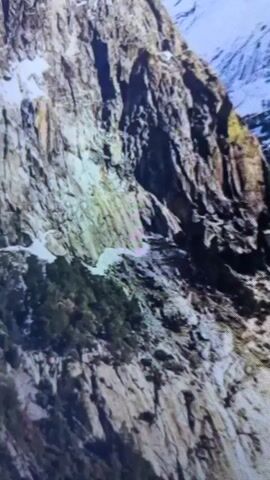 The video shows a panoramic view of a snow-covered mountain range. The mountains are rugged and appear to be made of granite. Snow is visible on the peaks and slopes, with some areas of exposed rock. The sky is blue with scattered clouds. The video is a still shot and does not contain any people, objects, or actions.