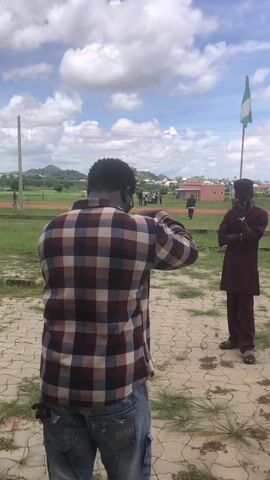 The video shows two men outdoors under a cloudy sky. The man in the foreground is seen from behind, wearing a plaid shirt and blue jeans. He is holding what appears to be a phone or camera in his hands, directed towards another man. This second man, standing to the right, is wearing a maroon outfit consisting of a tunic and trousers, and a matching cap. He has his arms crossed and is looking towards the man in the foreground. Behind the man in maroon, a Nigerian flag is flying on a flagpole. In the background, there are some buildings, a green field, and other people walking. The ground in the foreground is paved with interlocking bricks.