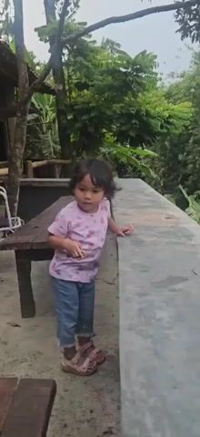 A young child with dark hair, wearing a pink t-shirt with a floral pattern and blue jeans, stands on a concrete patio. The child is looking down at a wooden table in front of them, with their hands resting on the table. In the background, there is a rustic building constructed with bamboo, a white chair with cushions, and lush green foliage. The camera pans slightly to the left and then to the right, showing the child interacting with the table and then looking towards the camera. Another person's hand briefly enters the frame from the left, touching the child. The child then turns and walks a short distance away, looking back over their shoulder. The video concludes with the child standing near some plants, still looking towards the camera.
