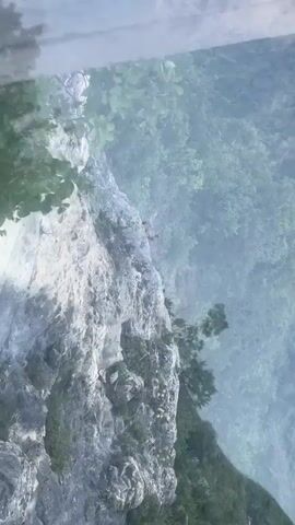 This is a vertical, eye-level, first-person perspective video of a glass-bottomed walkway over a steep, rocky cliff face covered in lush green foliage. The walkway is made of transparent panels, and the viewer is looking down at the precipice below. The cliff drops sharply into a densely wooded valley. The sky appears overcast or hazy, creating a soft, diffused light. The video appears to be filmed in a natural, mountainous environment, likely a tourist attraction. There are no other people visible in the frame. The overall impression is one of height and a sense of looking down from a precarious position.