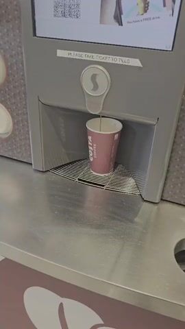 A coffee machine dispenses a drink into a paper cup. The cup is pink with the Costa Coffee logo. The machine is gray and has a screen with instructions. The scene is indoors, possibly in a cafe or supermarket.