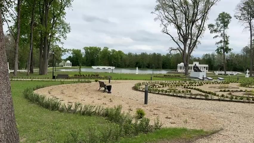 The camera captures a still, wide shot of a park or garden setting on a cloudy day. In the foreground, there is a winding path covered in light-colored gravel. Along the edges of the path are low-lying green bushes. Scattered across the gravel are small, newly planted shrubs, arranged in decorative circular patterns.  

To the left, a dark-colored park bench sits on the edge of the path, next to a grassy area with scattered trees. In the background is a tranquil pond or lake, with two white gazebo-like structures visible on the opposite shore. A white bridge spans the water between the two structures, and a fountain can be seen in the middle of the pond.

To the right of the gravel path, there is a neatly maintained lawn with several tall trees.  A white statue and another park bench are also visible in the background. The scene is peaceful and well-maintained, suggesting a serene and contemplative atmosphere.