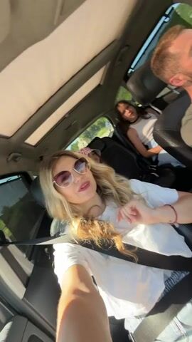 A blonde woman with sunglasses, wearing a white t-shirt, is filming a selfie video in a car. She is in the front passenger seat and has a seatbelt on. In the driver's seat, a man is visible. In the back seat, two girls are visible. One girl in a white top is behind the driver, and another girl in a patterned hat is behind the blonde woman. The car has a sunroof, and the sun is shining brightly. The blonde woman is singing along to music and dancing.
