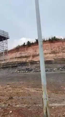 The video shows a wide, barren landscape that appears to be a quarry or mining site. A tall water tower stands prominently on the left, constructed with a metal frame and a large, square tank at the top. Behind it, a steep, layered cliff face reveals the earth's different strata with varying colors. 
The ground in the foreground is dusty and uneven, with patches of gravel and scattered debris. The sky is overcast with heavy clouds, suggesting a dull or potentially rainy day. 
As the camera pans to the right, several tall light poles become visible, lining a road that cuts through the landscape. In the distance, the land slopes upwards, forming hills covered with sparse vegetation. The scene conveys a sense of industrial activity and environmental impact.