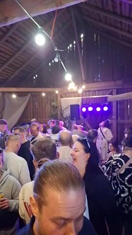 The video shows a crowded indoor gathering, likely a party or event, taking place in a wooden structure resembling a barn or hall. The ceiling is made of wooden beams, and strings of lights are strung across it, illuminating the space. Several people of various ages and genders are visible, mostly facing the camera or interacting with each other. Some are dancing or appearing to enjoy themselves. In the background, there is a stage area with lighting equipment, including purple and green lights. There are speakers and other electronic equipment on the stage. The lighting is dynamic, with colors changing, creating a festive atmosphere. The camera is held by someone in the foreground, and the focus shifts as the video progresses. Some individuals are laughing and talking, while others seem to be observing their surroundings. The overall impression is of a lively social event.
