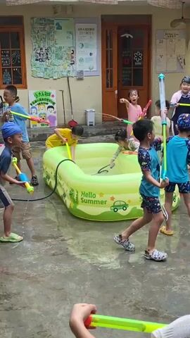 A group of children are playing in an outdoor courtyard, possibly a school or daycare. The ground is wet, suggesting recent rain or water play. In the center of the scene is a large, inflatable green kiddie pool with "Hello Summer" written on it. Several children are inside or around the pool, holding water guns and spraying water at each other. In the background, there is a building with windows and doors, adorned with colorful drawings and posters. Another group of children are standing on the paved area surrounding the pool, also holding water guns and participating in the water fight. The children are wearing casual summer clothing, and some are barefoot or wearing sandals. The atmosphere appears lively and joyful, with the children actively engaged in their water play.