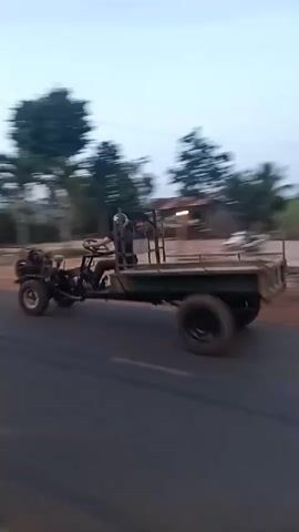 The video shows a vehicle moving along a road. A small, flat-bed trailer with large tires is being pulled by another vehicle ahead. Two people on scooters pass the larger vehicle that is filming. The landscape shows houses and trees along the roadside. The sky is overcast.