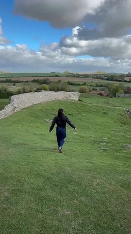 A daytime video captures a person, viewed from behind, walking down a grassy slope within an ancient ruin site. The person, dressed in a dark jacket and blue jeans, initially has their arms outstretched to the sides as they move away from the camera, then brings them closer to their body. To the left and right, stone walls, likely remnants of a historical structure, frame the grassy area. Beyond the ruins, a vast rural landscape unfolds, characterized by agricultural fields, scattered trees, and distant settlements under a partly cloudy sky with patches of blue. The overall setting is an expansive, open-air historical site in a countryside environment, consistent with locations in King's Lynn and West Norfolk, United Kingdom.