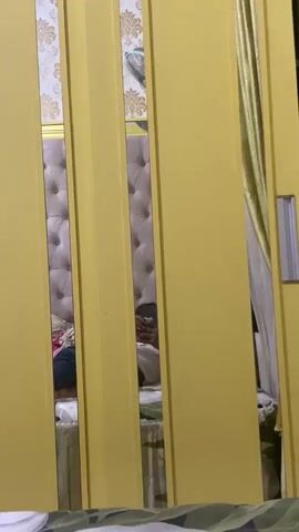 The video shows a reflection in a yellow sliding closet door. The reflection reveals a person holding a phone in their hands, seemingly taking a selfie. The background of the reflection shows a tufted headboard of a bed, suggesting the scene is set in a bedroom. There is also a glimpse of a bedside table with some items on it. The person's face is not clearly visible, but their hands and the phone are. The closet door has vertical panels, with the reflection appearing in the gaps between them.