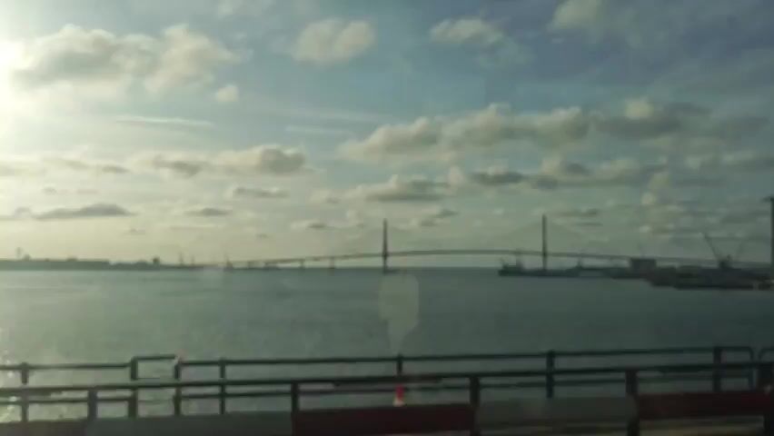 This video captures a scenic view of a long, cable-stayed bridge spanning across a wide body of water. The bridge has two prominent pylons that support its deck, with cables extending outwards to hold it in place. In the distance, a city skyline is visible, characterized by numerous buildings and structures. The scene also includes industrial elements such as cranes and ships, suggesting the presence of a port or harbor.

The time of day appears to be either early morning or late afternoon, as indicated by the soft, warm sunlight casting long shadows and illuminating the clouds. The sky is partly cloudy, with fluffy white clouds scattered across a pale blue expanse, creating a serene and slightly dramatic atmosphere. The water is calm, reflecting the sky and the bridge, adding to the tranquil mood.

The perspective suggests the footage is being filmed from a moving vehicle or a vantage point slightly elevated above the water, possibly from another bridge or a ferry. In the foreground, a metal railing with white and red markers runs horizontally, further reinforcing the idea of being on a transport or observation deck.

While there are no people explicitly visible, the presence of the infrastructure implies human activity and engineering prowess. The overall mood is peaceful and majestic, highlighting the grandeur of the bridge and its integration with the natural and urban landscape. The location is identified as Cádiz, Spain, a city known for its historical significance and coastal beauty, and this bridge is likely a significant landmark within the region. No visible text is present in the video.