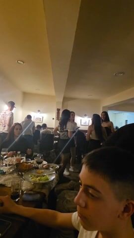 The video shows an indoor setting, likely a restaurant, bustling with activity. The camera moves to show a group of men seated around a table filled with food and drinks. A younger man stands between tables playing guitar and singing. Some of the seated men are talking and laughing. Several women stand off to the side.