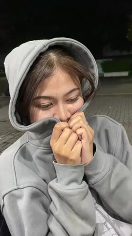 A young woman with brown hair, wearing a grey hoodie, peeks out from under the hood and smiles at the camera. Her hands are clasped in front of her face, holding onto the fabric of the hoodie. The background is dark and out of focus, with some lights and trees visible. The video then cuts to a blurry shot of a white car and then to two people sitting at a table outdoors at night. The person on the left is wearing a red and white patterned dress, and the person on the right is wearing a black t-shirt and a grey baseball cap. They are both smiling and looking at each other. There is a white cigarette pack and a smartphone on the table.