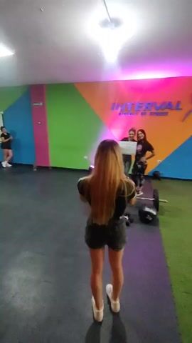 Here's a description of the video:

The video shows the interior of a fitness studio with brightly colored walls. In the background, a group of people are standing, possibly observing. The focal point is on a woman with long blonde hair, wearing a black top and dark shorts, standing facing away from the camera. In front of her, two other people appear to be posing for a photo with a certificate or award. There is a barbell with weights nearby. The floor is a mix of gray rubber and green artificial turf. A wooden plyometric box is visible on the right.