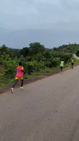 The video shows three people running on an asphalt road. All three are wearing athletic clothing. The first person has a yellow shirt, pink vest with the number 8 on it, black shorts, and white sneakers. The second person is wearing a yellow-green tank top with the number 15 on it, dark blue shorts, and white sneakers. The third person has a yellow-green tank top with the number 13 on it, dark blue shorts, and black sneakers. The asphalt road curves uphill in the background. The landscape is green and lush. The sky is cloudy.