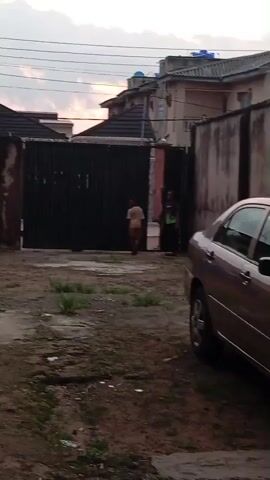A group of children and adults are gathered near a large black gate. The children appear to be playing or interacting with each other, while the adults seem to be supervising or involved in a conversation. A silver car is parked to the right of the frame, and houses are visible in the background. The sky is cloudy, suggesting it might be evening or early morning.