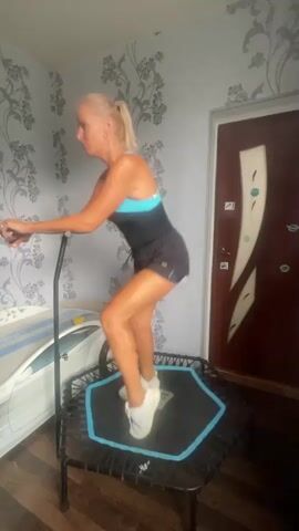 A blonde woman in athletic wear is performing a squatting motion on a hexagonal trampoline. The trampoline has a blue and black frame and mat. The woman is holding onto a black handle for balance. She is wearing a black and blue one-piece swimsuit with shorts, white athletic shoes, and her hair is pulled back in a ponytail. She is in a room with floral wallpaper, a bed with a striped comforter on the left, and a wooden door on the right. The lighting appears to be natural light from a window or overhead. The video is composed of a series of still images, creating a stop-motion effect.