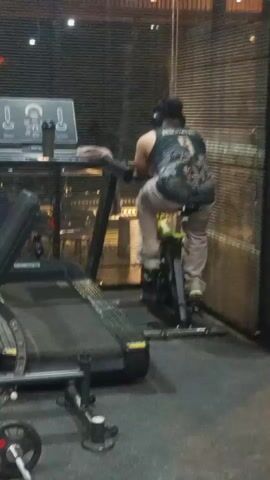 A person wearing headphones and a dark sleeveless shirt with the number 9 on the back is riding an exercise bike in a gym. The person is wearing light-colored pants. Next to the exercise bike is a treadmill. In the foreground, there is gym equipment, including weights. The environment appears to be indoors with wooden blinds on a wall in the background.
