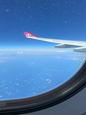 The video shows a view from an airplane window, featuring the plane's wing with the Turkish Airlines logo against a backdrop of blue sky and scattered clouds. The window is somewhat dirty with spots. The view includes the horizon where the sky meets the ocean or land.