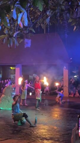 The video shows a fire dancing performance at night with an audience in the background. Four performers are visible, three are standing and one is kneeling on the stage. All four performers are wearing colorful clothing. They manipulate fire props, including torches and spitting fire. The stage is wet, reflecting the light from the fire. A rock structure with a face carved into it stands on the left side of the stage.  Above the stage are branches of trees, lit up by stage lighting. The lighting shifts between orange, blue, and purple. Some audience members can be seen holding up their phones, presumably to record the performance.