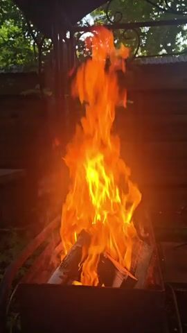 A close-up shot shows a fire burning in a metal grill. The flames are bright orange and yellow and reach upwards. Logs are visible in the grill. The background is out of focus and shows trees and a wooden fence. The scene appears to be outdoors.