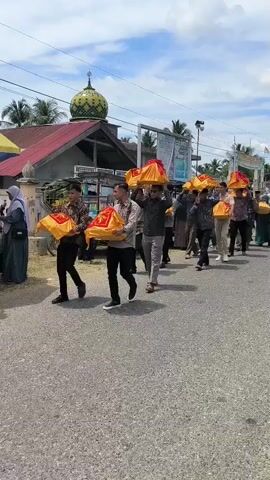 A group of men are walking in a procession down a paved road. They are carrying objects wrapped in red and gold fabric over their heads. Some of the men are wearing traditional attire, while others are in more casual clothing. Behind them, a mosque with a green dome is visible. To the left of the road, there is a colorful umbrella and a stand that appears to be selling food or drinks. Further down the road, more people and tents are present, suggesting a local event or gathering. The sky is overcast with some visible clouds.