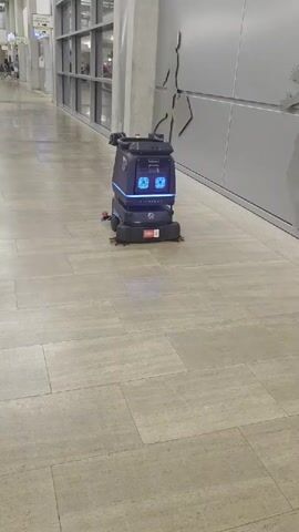 A robot cleaner with a blue illuminated display moves across a tiled floor in a hallway. The display on the robot shows text. The robot has brushes on the bottom and a camera or sensor on top. The hallway has a grey wall with a graphic on it and a tiled floor.