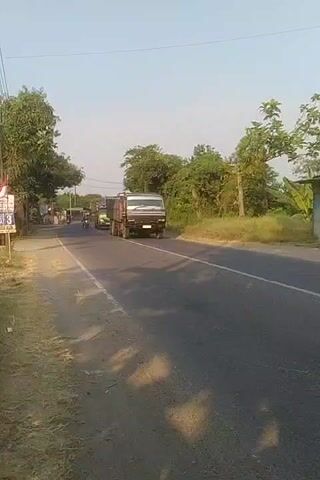 The video shows a road with several vehicles. In the foreground, on the right side of the road, a large dark-colored truck with a metal cargo bed is driving away from the camera. The road is paved and has a white line marking the center. On the left side of the road, there is a dirt shoulder with some trees. Further down the road, there are more vehicles, including a green dump truck with a red cargo bed, a smaller red truck, and several motorcycles. People are visible riding the motorcycles. The environment appears to be rural, with trees and vegetation lining the road. The sky is clear and blue, suggesting daytime. The camera is mounted low and appears to be moving with the flow of traffic, possibly on a motorcycle or in a vehicle behind the main subjects.